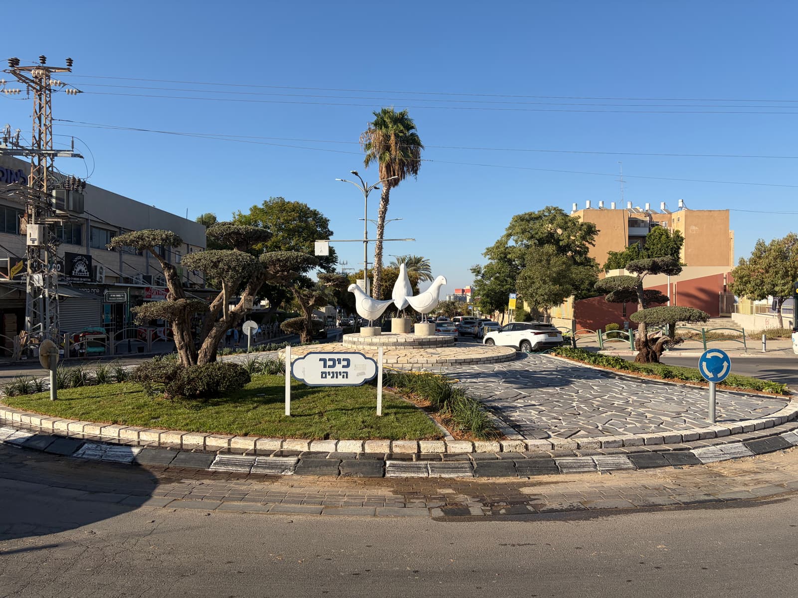 כיכר היונים באופקים — שלוש יונים לבנות על במה מרוצפת, שלט הכיכר וגינון חדש על ציר שדרות הרצל