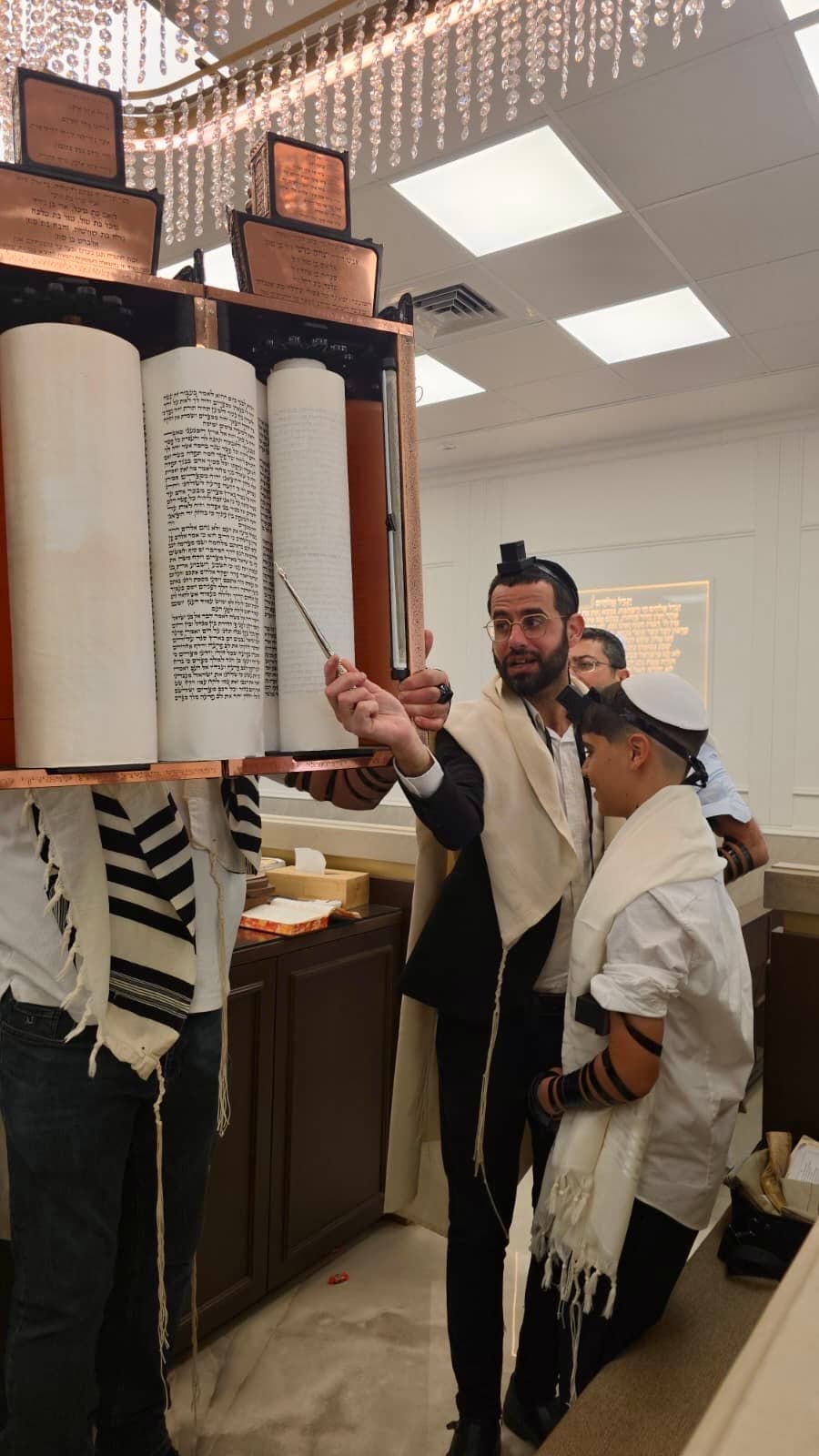 ינון מצליח מצביע עם יד הכסף על ספר התורה לצד חתן בר מצווה