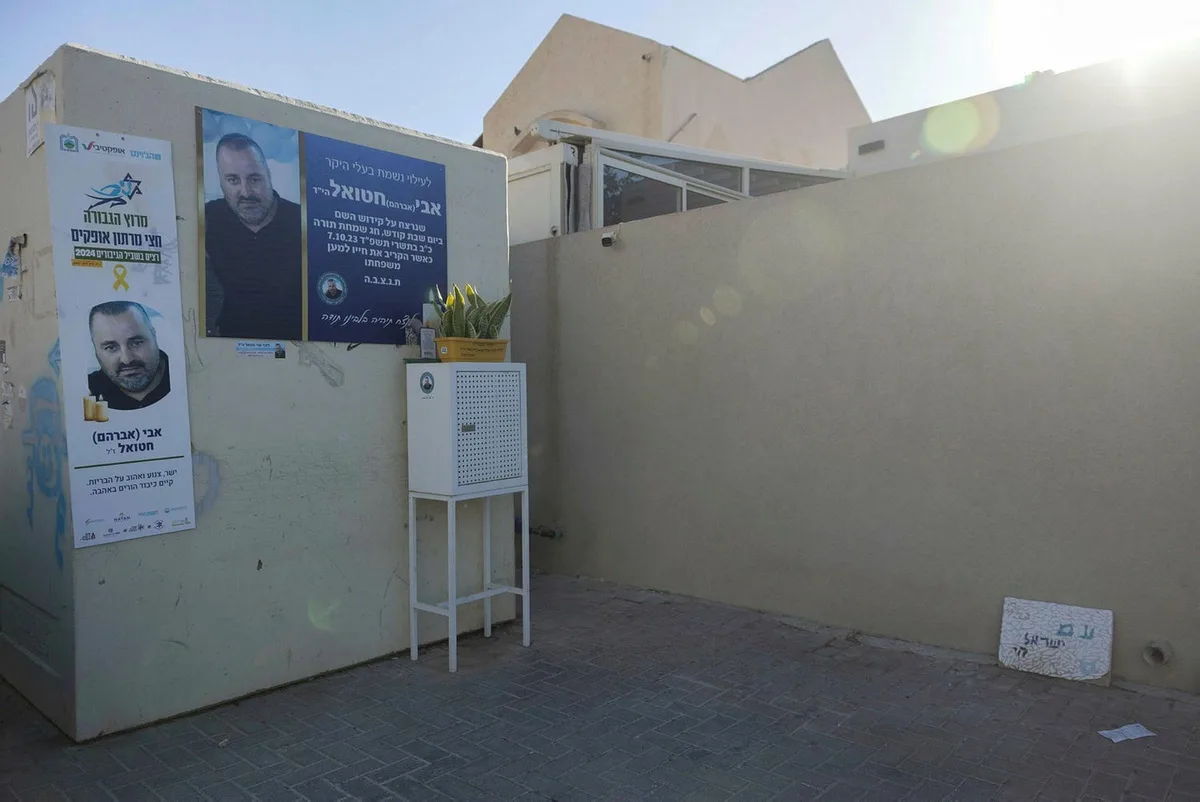 פינת הנצחה לאבי חטואל הי״ד במישור הגפן: שלט זיכרון על הקיר וארון נרות נשמה לבן על המדרכה