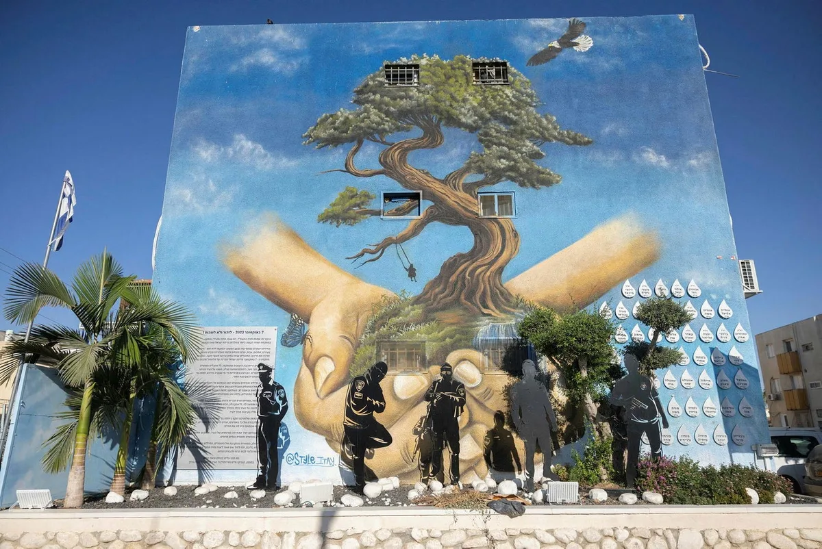 ציור הקיר במישור הגפן באופקים — עץ הנשען על כפות ידיים, צלליות השוטרים שהגנו על השכונה וקיר הטיפות עם שמות הנרצחים